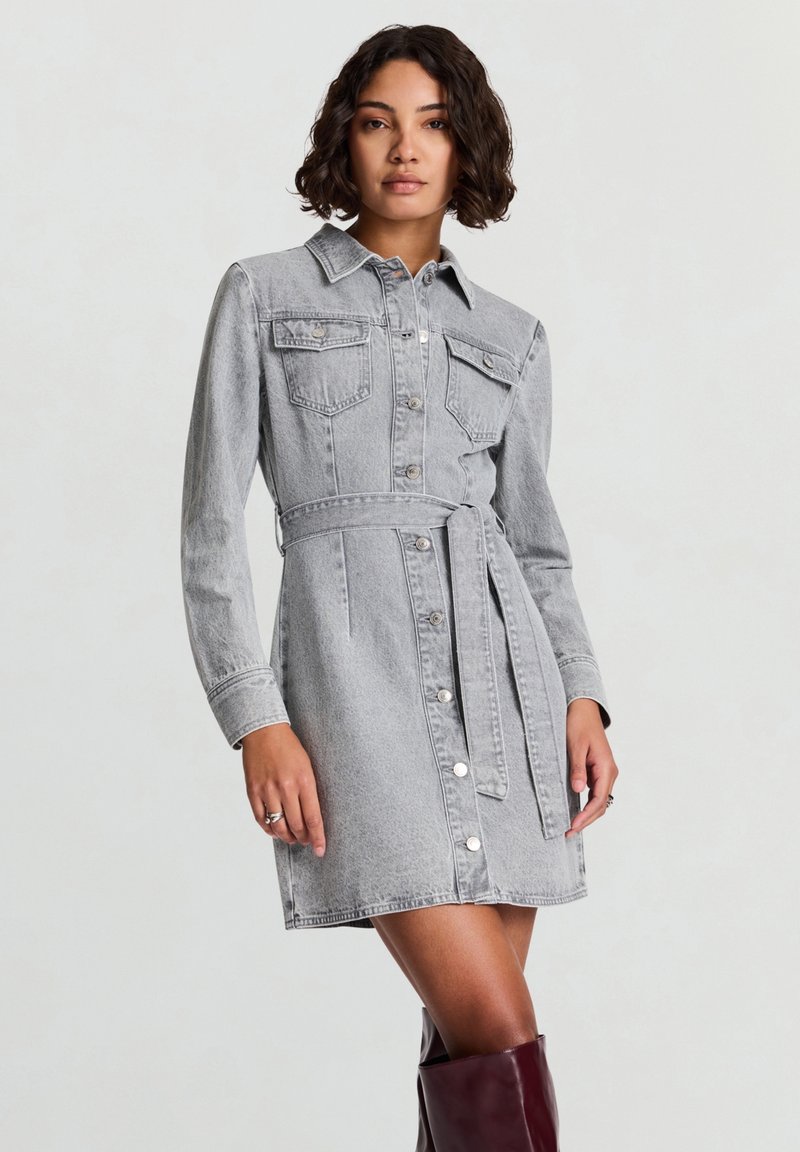 Vestito camicia in denim grigio con maniche lunghe, davanti con bottoni, collo appuntito, due tasche sul petto e cintura abbinata. Texture liscia, vestibilità strutturata.