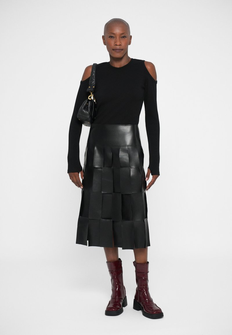 Vrouw met een geschoren hoofd, gekleed in een zwarte cold-shoulder top, een knie-lange leren rok met patchwork, bordeauxrode laarzen en een zwarte schoudertas.