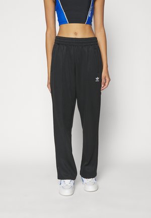 Kvinde iført sorte Adidas træningsbukser, hvide sneakers og en sort og blå cropped tanktop, der står mod en ensartet baggrund.
