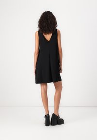 Robe noire sans manches avec un dos en V, présentant une coupe évasée et une texture lisse, accompagnée de chaussures à talons épais noires.