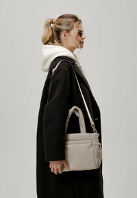 Beige laukku, jossa on tikattu pinta, lyhyet kantokahvat ja pitkä säädettävä hihna. Laukussa on etutasku ja sileä tekstuuri.