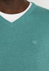 Turkusowy sweter dzianinowy z dekoltem w serek, z białym wewnętrznym kołnierzykiem i małym haftowanym logo na piersi.