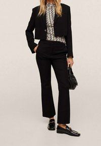 Blazer negro corto sobre una blusa estampada, combinado con pantalones acampanados negros y mocasines brillantes. El atuendo incluye un bolso de cuero.
