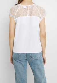 Weißes Oberteil mit Spitzenrücken und kurzen Spitzenärmeln. Strukturierte Frontpartie, Knopfverschluss am Hals, kombiniert mit Jeans.