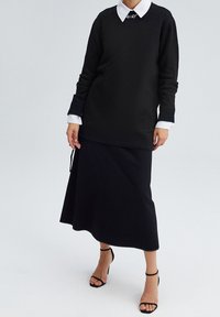 Maglione nero con scollatura rotonda e maniche lunghe indossato su una gonna nera. L'outfit presenta una camicia bianca con colletto sotto.