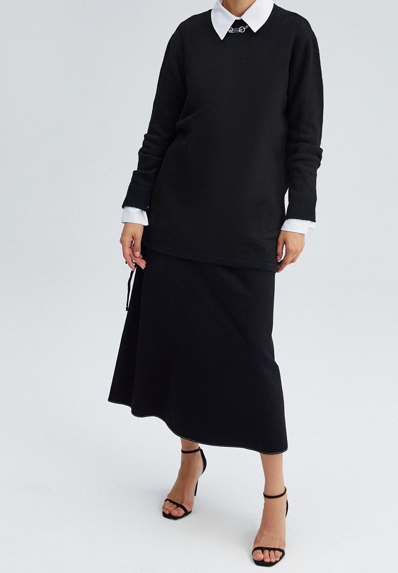 Maglione nero con scollatura rotonda e maniche lunghe indossato su una gonna nera. L'outfit presenta una camicia bianca con colletto sotto.