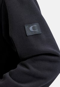Schwarzes Sweatshirt mit einer weichen Textur und figurbeduldigem Design, ausgestattet mit einem quadratischen schwarzen Patch mit einem grauen Logo auf dem linken Ärmel.