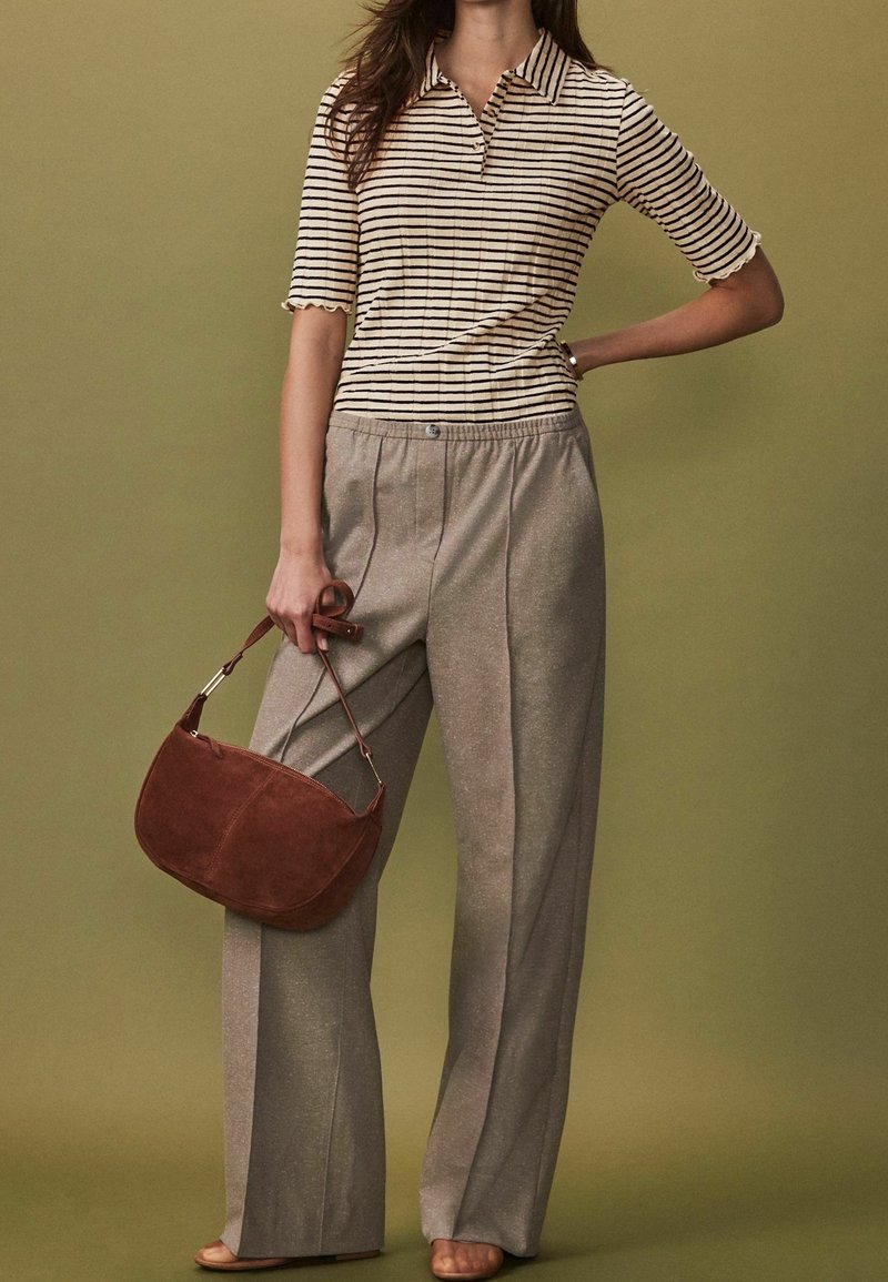 Femme portant un pantalon large beige et un haut rayé crème à manches courtes, tenant un sac à main en cuir marron sur un fond olive.
