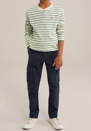 Camisa de manga larga a rayas en verde claro y blanco con botones de madera, combinada con pantalones cargo navy con bolsillos laterales y zapatillas beige.