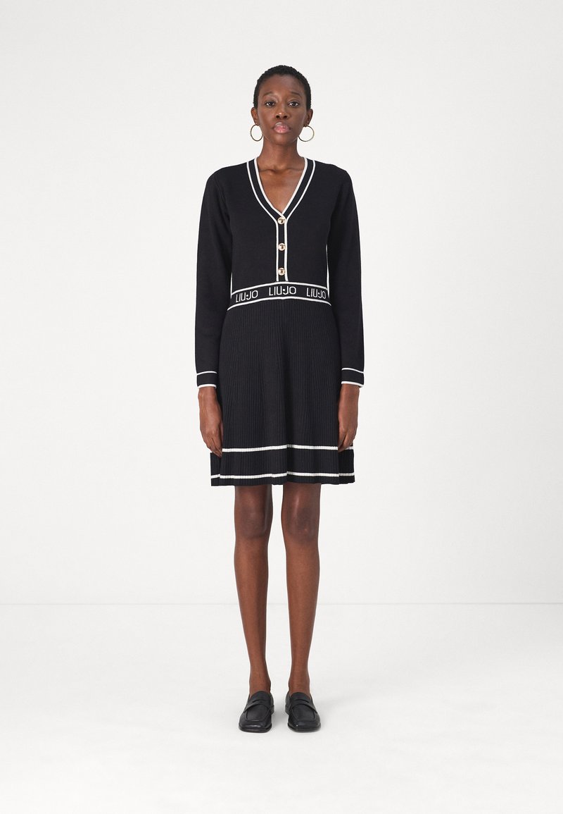 Robe noire en tricot avec manches longues, décolleté en V, boutons dorés et jupe côtelée. Présente des rayures blanches et une ceinture à logo. Associée à des chaussures noires.