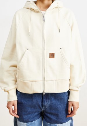 Person trägt einen cremefarbenen Kapuzenpullover mit Reißverschluss, Fronttaschen und braunem Logopatch, kombiniert mit blauen Jeanshosen mit Patchwork.