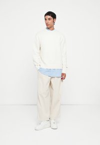 Fehér pamut pulóver egy kék csíkos ing fölött, off-white széles szárú nadrággal és fehér sportcipőkkel párosítva. Egyszerű, laza szabású.