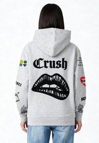 Grijze hoodie met een grote zwarte afbeelding van lippen op de achterkant, diverse patches en tekst op de mouwen. Comfortabel materiaal, casual ontwerp.