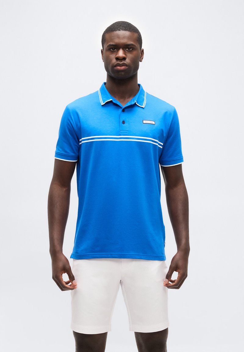 Άνδρας που στέκεται με χαλαρά τα χέρια, φοράει μπλε πόλο με λευκές ρίγες και λευκά σορτς, σε απλό φωτεινό φόντο.