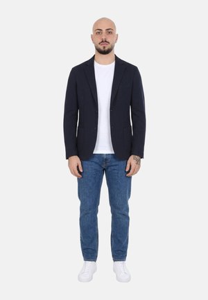 Uomo in piedi con espressione neutra che indossa un blazer nero, una t-shirt bianca, jeans blu e sneakers bianche su sfondo bianco.