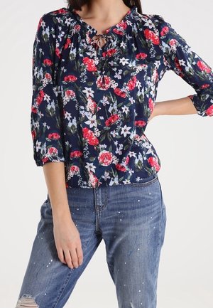 Žena nosící tmavě modrou květovanou halenku s 3/4 rukávy a vázáním u krku, v kombinaci s modrými džíny s opotřebovaným vzhledem a bílými skvrnami barvy.