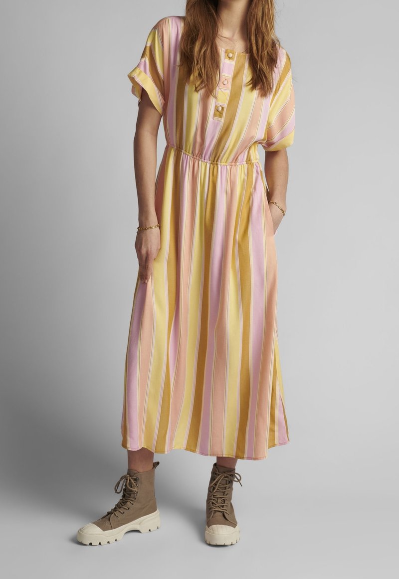 Robe maxi rayée rose et jaune, avec des boutons au niveau de l'encolure, une taille élastique et des manches courtes. Associée à des bottines marron à lacets.