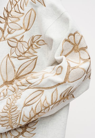 Μανίκι φούτερ με μπεζ κεντήματα από λουλούδια και φύλλα σε ανοιχτό γκρι ύφασμα, με εμφανείς λεπτομερείς ραφές και καμπύλες πτυχώσεις.