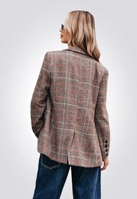 Blazer à carreaux en marron, rouge et gris avec une coupe ajustée, des revers crantés et des poignets boutonnés. Assorti à un jean bleu à jambes larges.