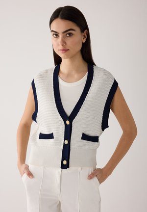 Jeune femme aux longs cheveux foncés portant un gilet en tricot blanc avec bordure marine et boutons dorés par-dessus un haut blanc, les mains dans les poches d'un pantalon blanc.