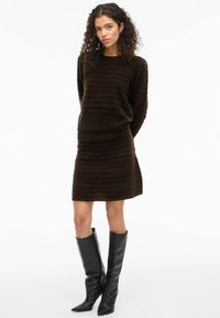 Donna con capelli ricci, che indossa un vestito a maglia strutturato color marrone scuro e stivali neri a punta fino al ginocchio, in piedi davanti a uno sfondo bianco.