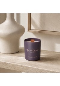 Bougie parfumée violette étiquetée Velvet Rose avec une mèche en bois sur une surface en pierre à côté d'un vase sculptural beige mat et une œuvre murale encadrée.