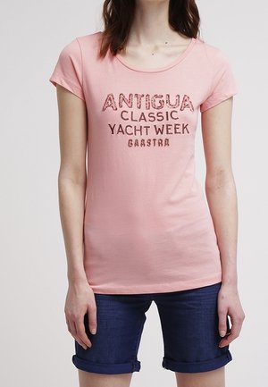 Różowa bawełniana koszulka z krótkim rękawem, z ciemnym nadrukiem "ANTIGUA CLASSIC YACHT WEEK". Noszona z dżinsowymi szortami.