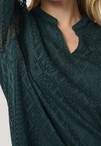 Haut en tissu texturé vert foncé présentant un motif géométrique, un col cranté et des détails en dentelle sur les manches.