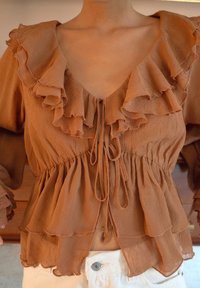 Bruine blouse met volants van textuurstof en striksluiting, gedragen met witte broek, met focus op details van de taille en de halslijn.