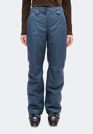 Modèle portant un pantalon de ski bleu avec une poche latérale zippée et des chaussures de ski noires.