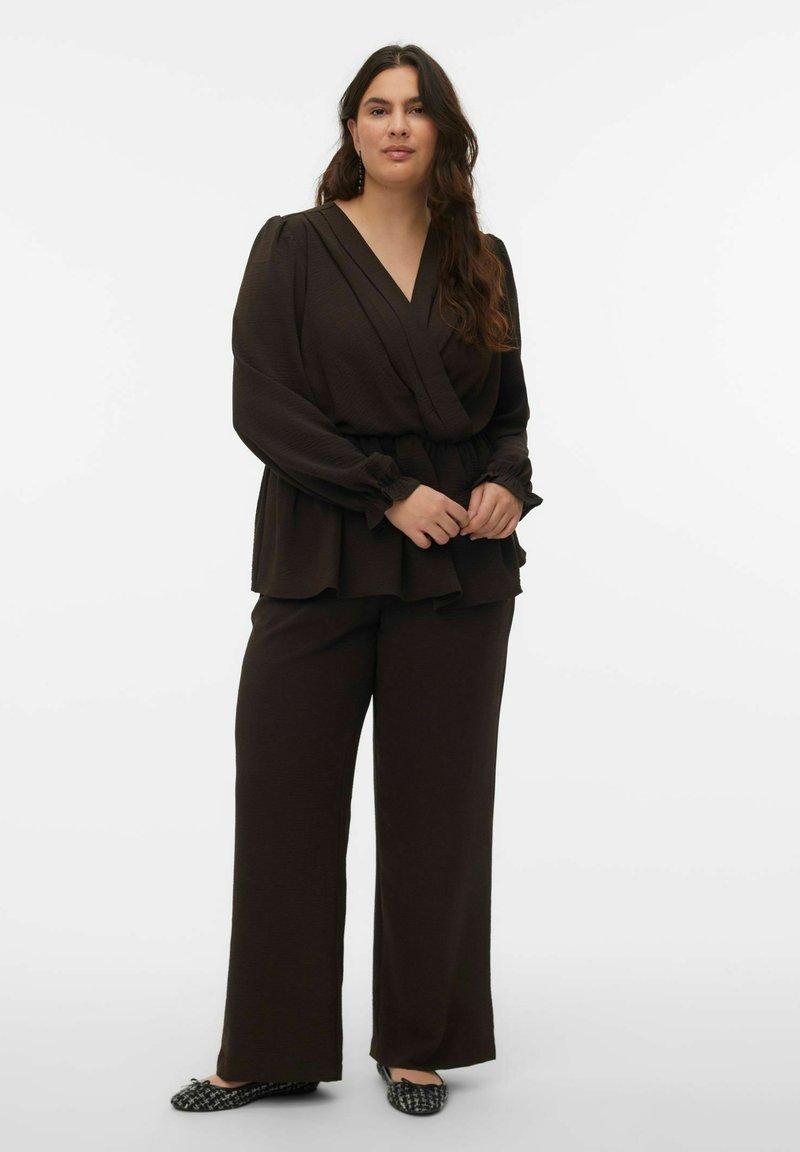 Rjava preklopna bluza z dolgimi rokavi in nagubani manšeti, kombinirana z rjavimi hlačami širokih nog. Obutev ima teksturiran črn vzorec.