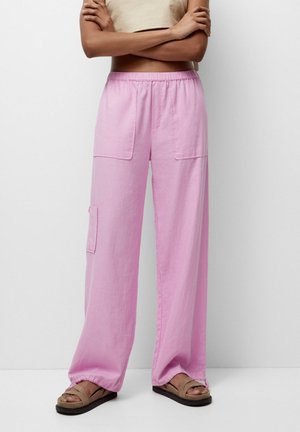 Femme portant un pantalon cargo rose ample avec de grandes poches avant et des sandales beiges, debout, les bras croisés.