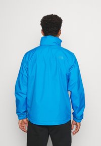 Giacca leggera blu, materiale resistente all'acqua, colletto alto, polsini elasticizzati, logo sul retro, texture liscia, progettata per l'uso all'aperto.
