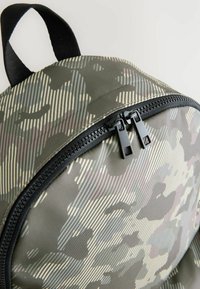 Rucksack mit einem Tarnmuster, ausgestattet mit einem strukturierten Stoff, schwarzen Reißverschlüssen und einem oberen Griff zum Tragen.