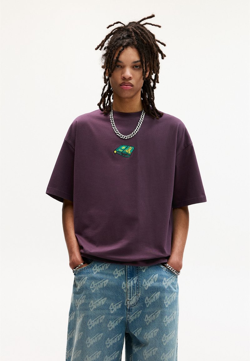 Overdimensioneret lilla t-shirt med et lille grønt og gult grafik på brystet, parret med lyseblå jeans med et gentaget hvidt logo-mønster.