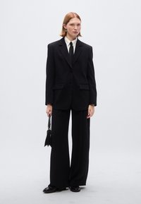 Μαύρο υπερμεγέθες σακάκι με δομημένους ώμους και πέτο με σχισμή, σε συνδυασμό με παντελόνια με φαρδιά μπατζάκια. Λευκό πουκάμισο και μαύρη γραβάτα ολοκληρώνουν το σύνολο.