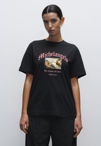 Sort bomulds T-shirt med grafik af Michelangelos "Skabelsen af Adam" med pink tekst ovenfor og nedenfor billedet. Afslappet pasform.
