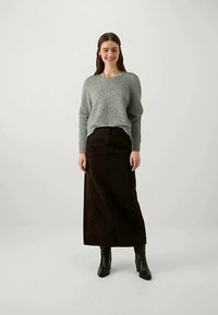 Sieviete, ģērbusies pelēkā džemperī, garā tumši brūnā svārku un melnās potīšu zābakos, stāv un smaida pret tīru baltu fona.