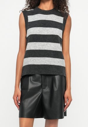 Žena v bezrukávovém svetru se šedými a černými vodorovnými pruhy a černých kožených šortkách stojí před čistě bílým pozadím.