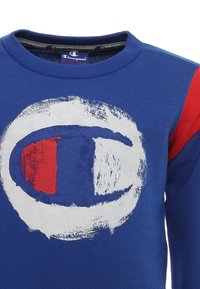 Modrá mikina s velkým bílým kruhovým logem, na kterém jsou červené a námořnické akcenty. Výstřih je ribbovaný a rukáv má červené detaily.