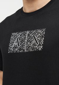 Čierne bavlnené tričko s obdĺžnikovou bielou grafikou so vzájomne prepletenými písmenami a vzormi na hrudi. Dizajn s okrúhlym výstrihom.