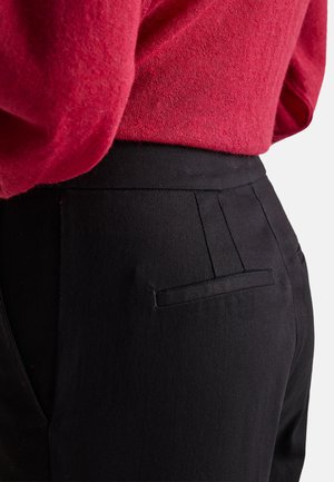 Zwarte broek met een gladde textuur, voorzien van een enkele achterzak en plooien, gedragen met een rode trui.