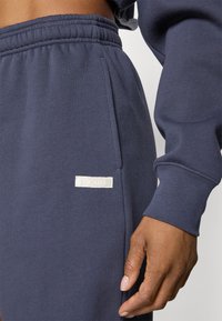 Calças de fato em azul-marinho com cintura elástica e bolsos laterais. Apresenta um pequeno rótulo retangular branco com "BORG" cosido na lateral.