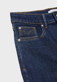 Jean en denim bleu foncé en gros plan montrant une poche avant avec des coutures orange et des rivets métalliques sur un fond clair.