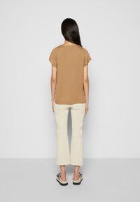 Braune Bluse mit kurzen Ärmeln und lockerer Passform, kombiniert mit cremefarbenen Schlaghosen und schwarzen Sandalen mit weißer Sohle, von hinten betrachtet.