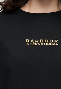 Μαύρο μπλουζάκι με μπεζ κεντημένο κείμενο "BARBOUR INTERNATIONAL" στο πάνω μέρος του στήθους, φορεμένο από άτομο με σκούρα μαλλιά.