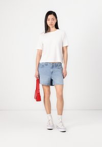 T-shirt blanc court, short en denim bleu clair, sac à main rouge, baskets montantes blanches et chaussettes blanches, centré sur un fond uni.