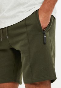 Homme portant un short vert olive avec une poche zippée et un cordon de serrage, la main dans la poche, assorti à une chemise gris clair.
