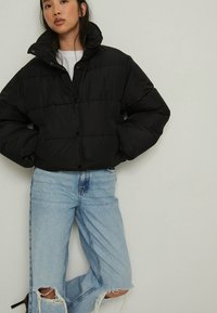 Melna pūkainā jaka ar augstu apkakli, pogu aizdari un iedobeņu dizainu. Saskaņota ar gaismas zila nolietotiem džinsiem ar saplēstiem malām.