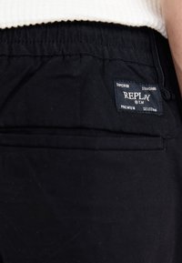 Svarta tygbyxor med elastisk midja, med en synlig etikett som visar "REPLAY" och ytterligare text om kvalitet och urval.